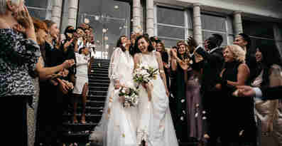 Två kvinnor i bröllopskläder går ner för en trappa där deras bröllopsgäster står och firar dem. 