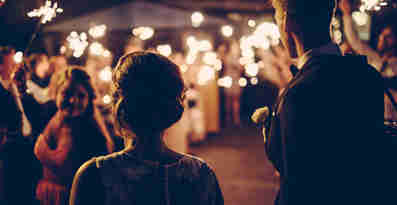 Ett bröllopspar står med ryggen mot kamera, framför sina bröllopsgäster som håller i tomtebloss som lyser i mörkret.